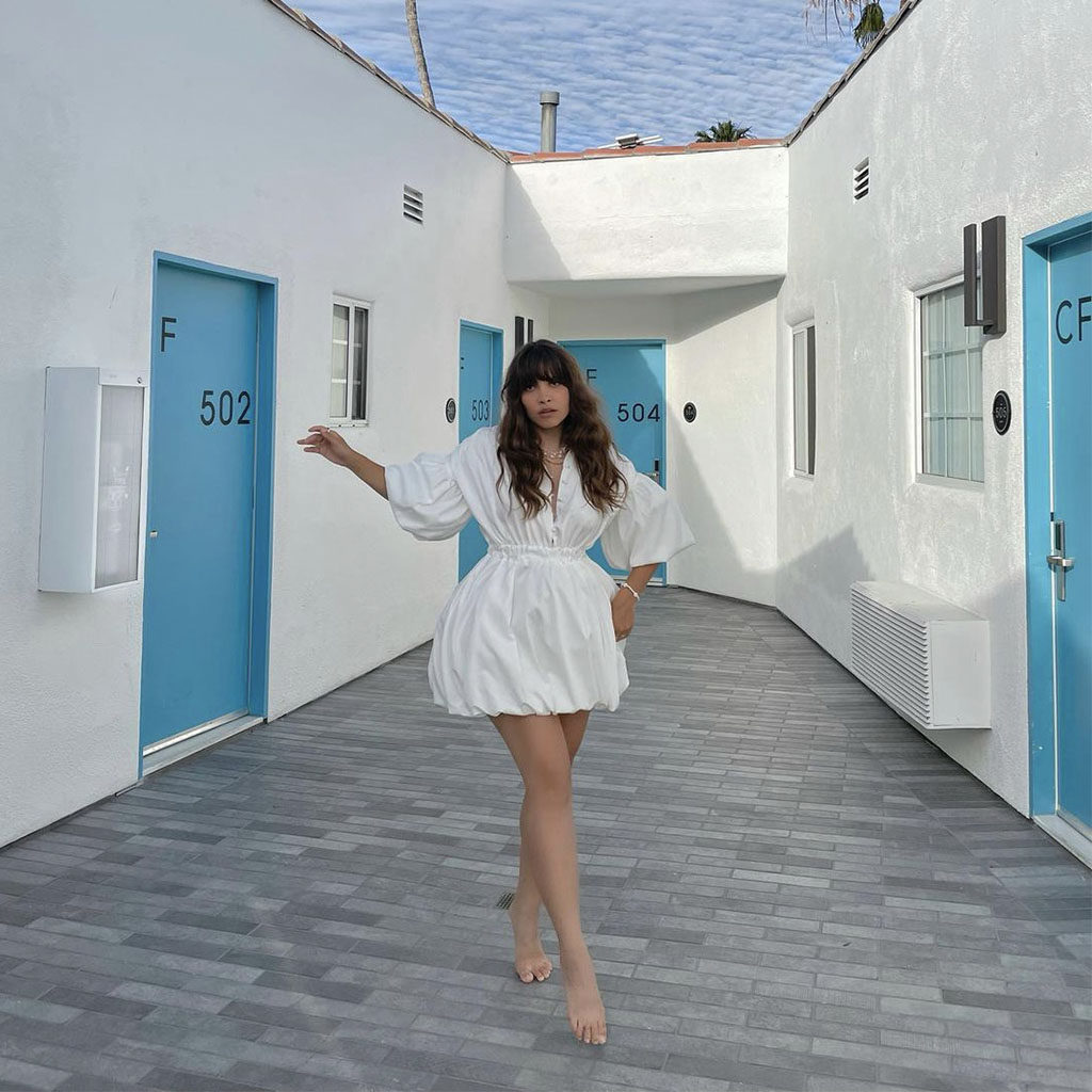 Woman in a white dress standing on her tippy toes posing in the stark white, outdoor hallway with blue doors of Bellanca Hotel
