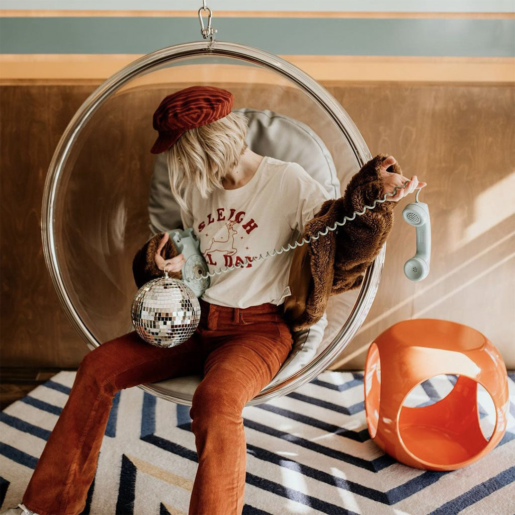 Woman holding a disco ball and old school telephone sitting in a hanging bubble chair in the lobby of the Rambler Motel