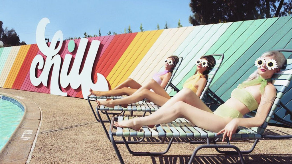 Three women wearing daisy sunglasses lying in sun loungers in front of the CHILL sign in the pool area of the Rambler Motel