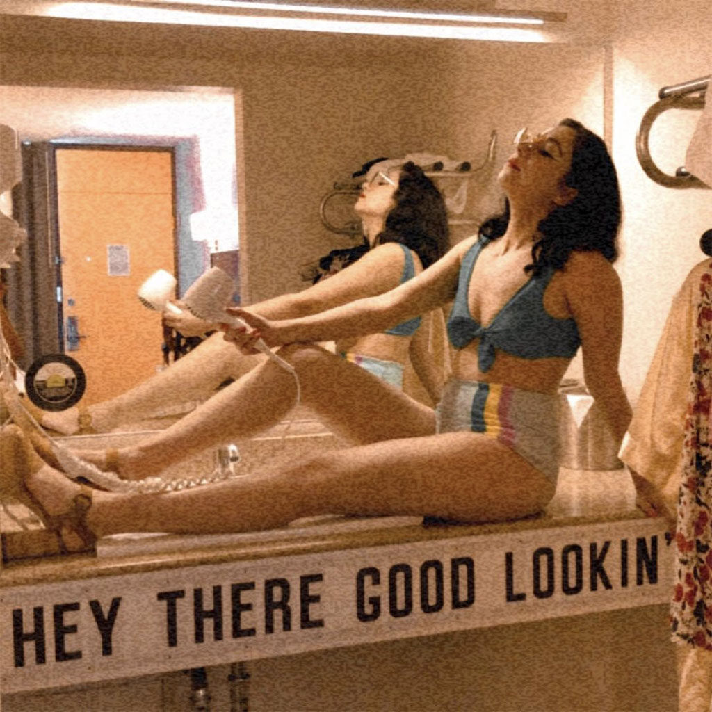 Woman holding a blow dryer sitting on top of the "Hey There, Good Looking" vanity in a Rambler Motel guestroom