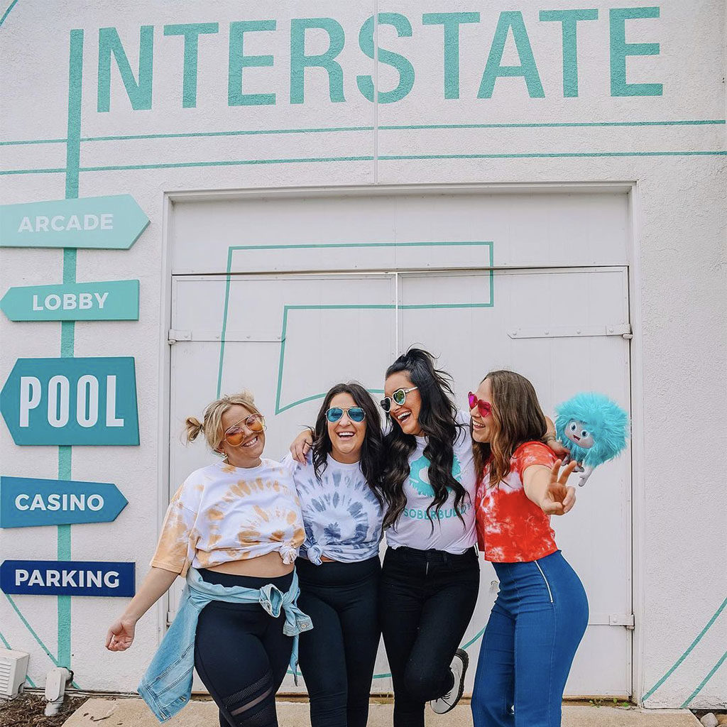 Four women smiling and hugging in front of the Interstate roadtrip styled directional signage at The Rambler Motel