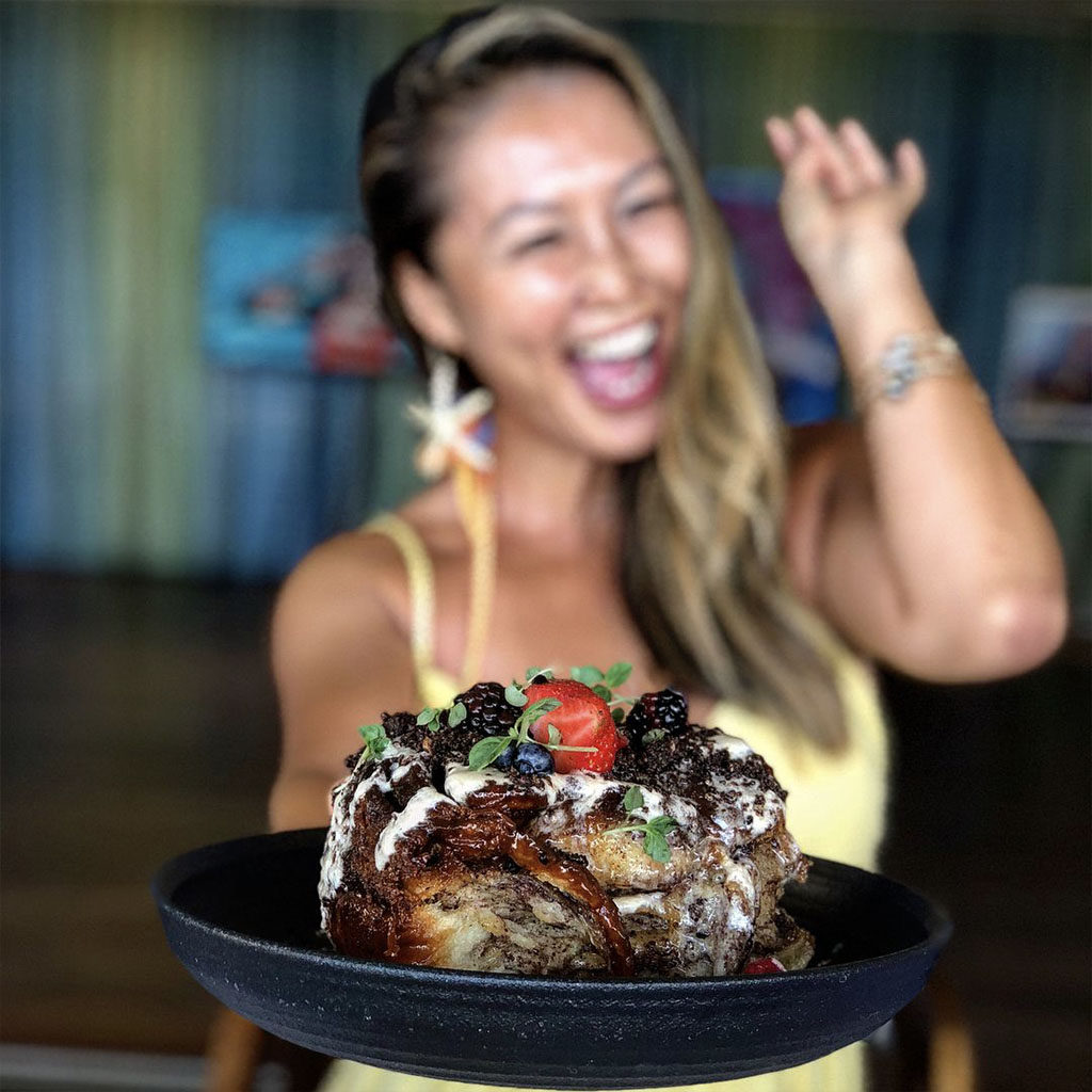 Woman smiling and holding a giant sticky bun topped with blackberries, strawberries, icing and chocolate cookie crumbles