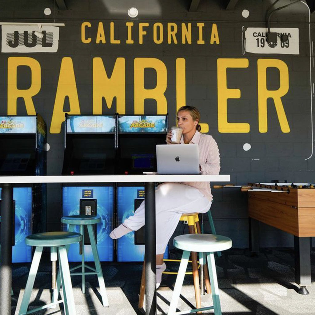 Woman sipping coffee at her laptop at a table in front of the Rambler license plate mural at Rambler Coffee Shop