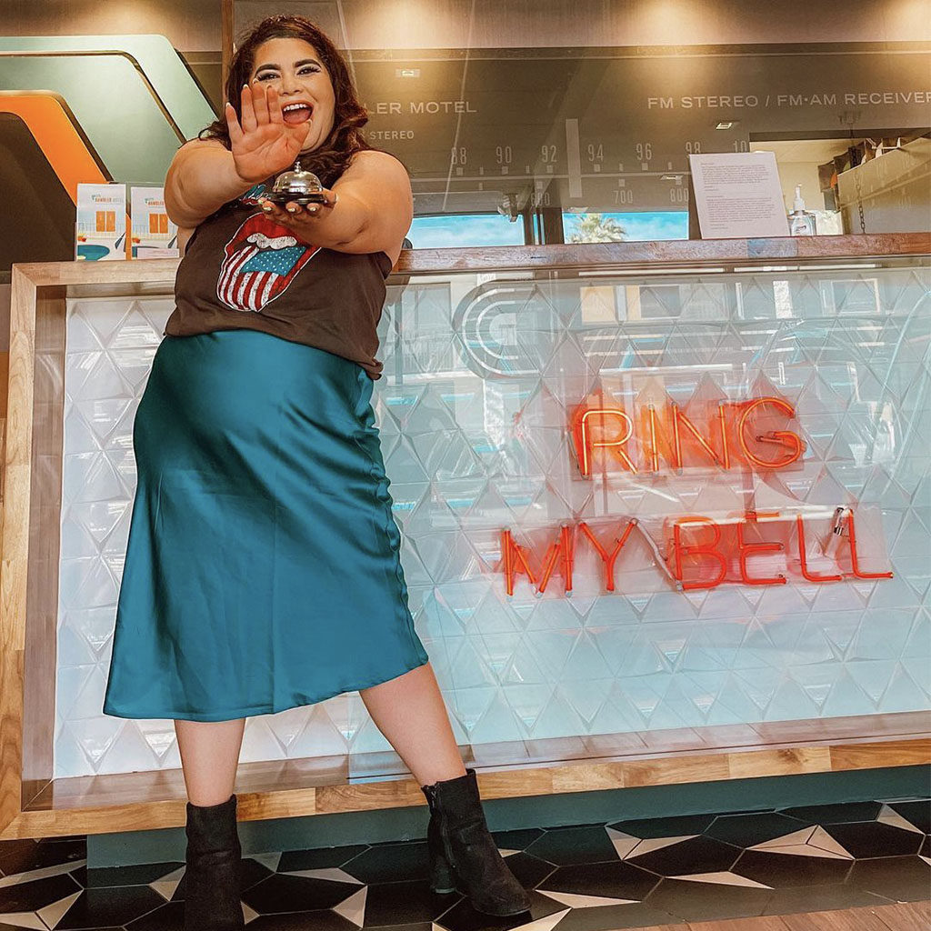 Woman smiling holding a bell in front of the "Ring My Bell" front desk check in at The Rambler Motel