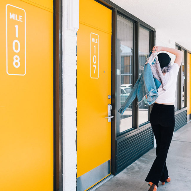 Girl walking with a jean jacket walking out two orange hotel room doors that say mile 108 and mile 107