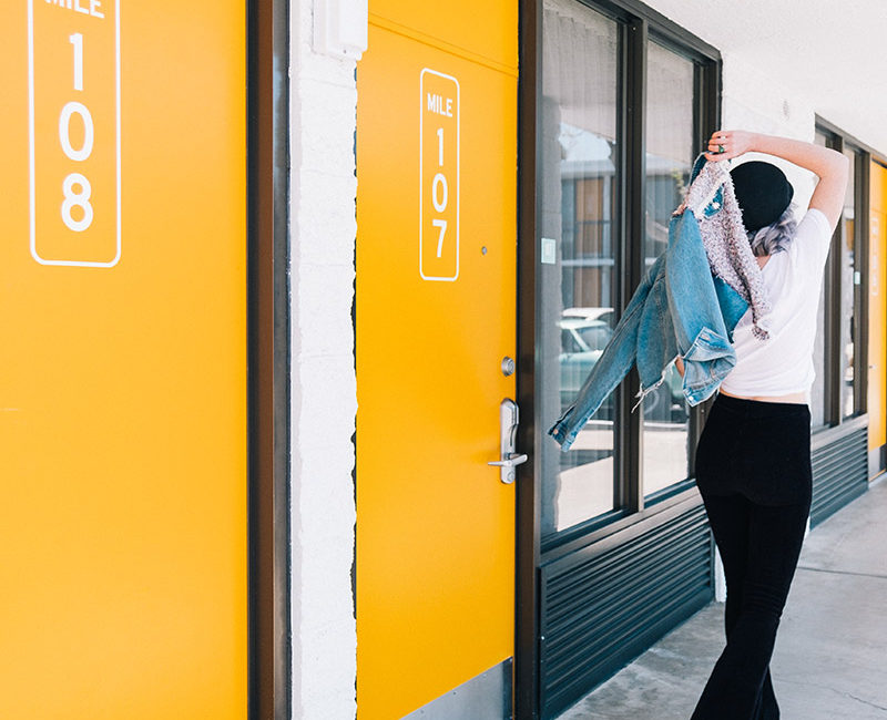 Girl walking with a jean jacket walking out two orange hotel room doors that say mile 108 and mile 107