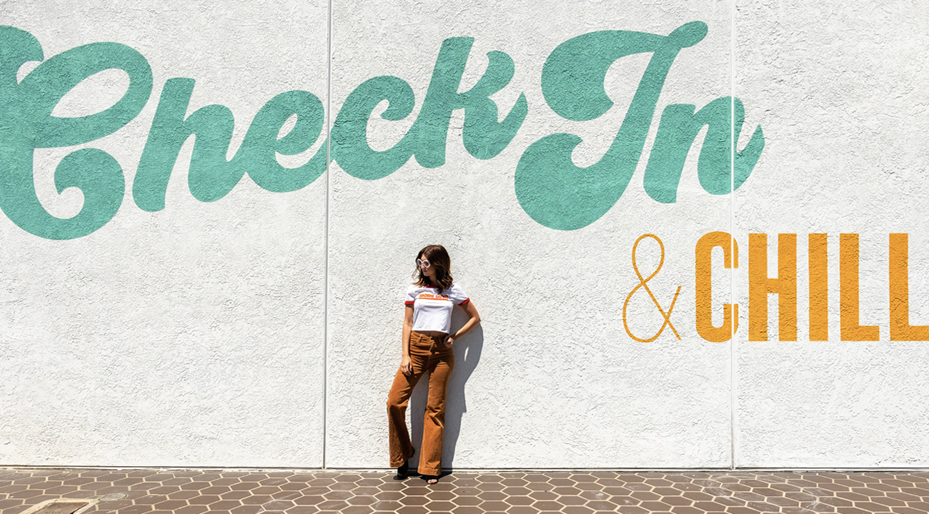Girl in tan bell bottoms standing in front of the Check In & Chill wall art at The Rambler Motel