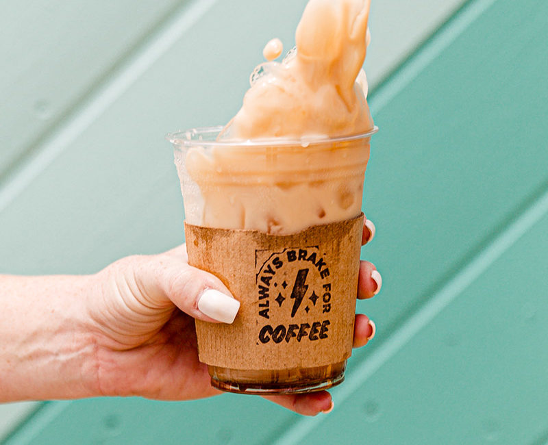 Hand holding an iced coffee that's spilling with a hand stamped sleeve that says "Always Brake for Coffee" against a light teal background