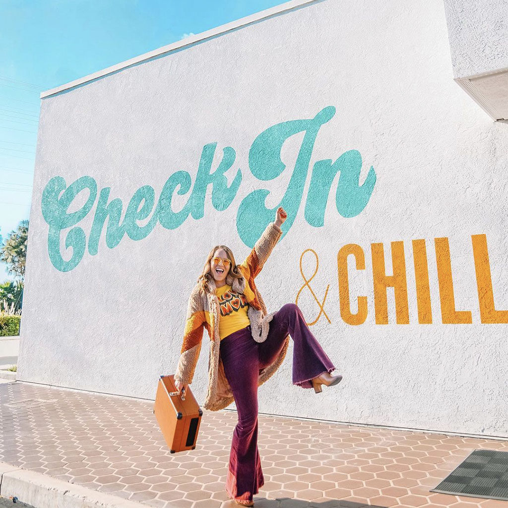 Woman holding a suitcase in bellbottoms posing in front of the "Check In & Chill" building mural at The Rambler Motel