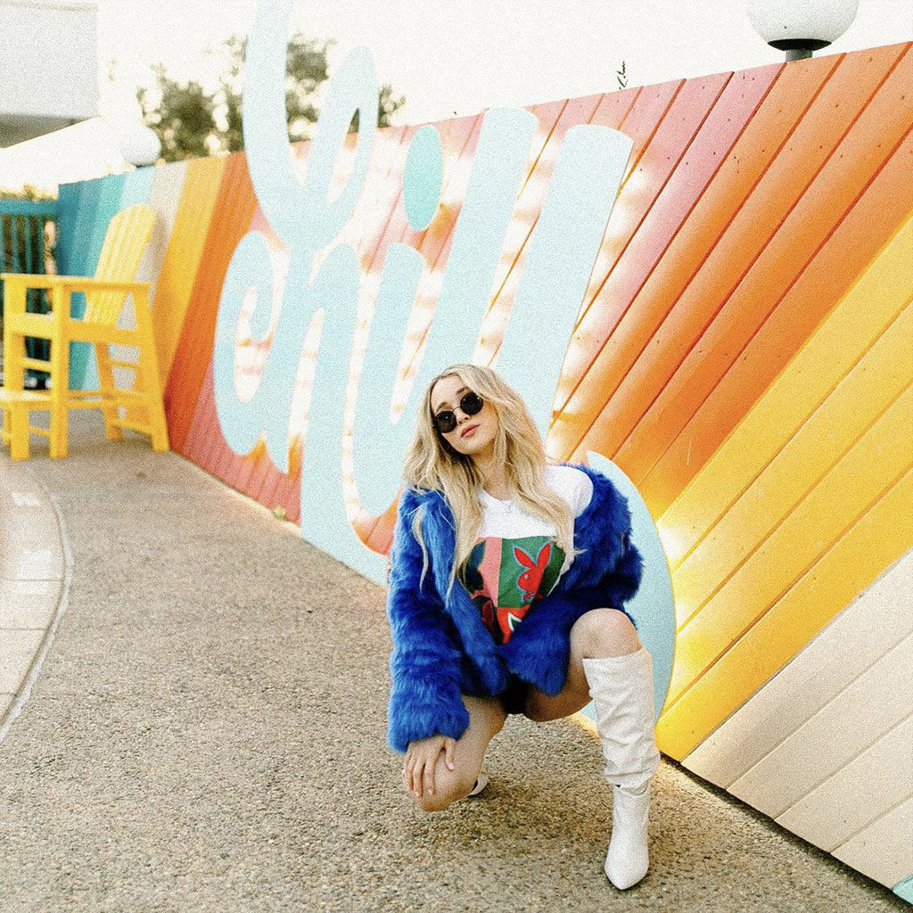 Woman in a blue furry coat and white boots kneeling in front of the CHILL sign at the pool of The Rambler Motel