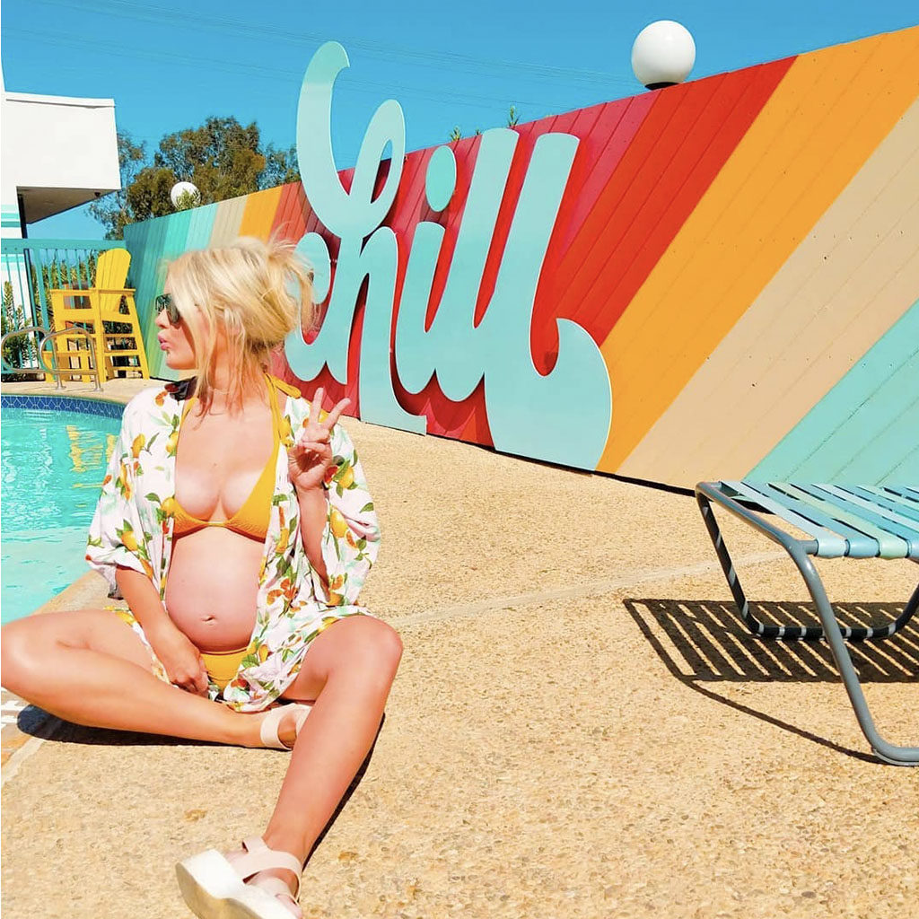 Woman in a yellow bathing suit throwing a peace sign sitting in front of the CHILL sign at the pool of The Rambler Motel