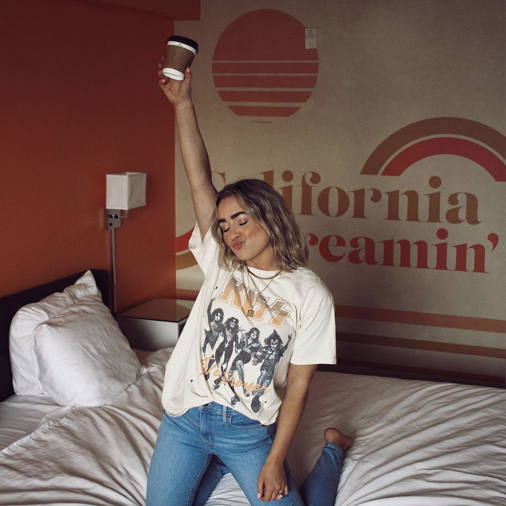 Women throwing up her coffee cup kneeling on the bed in front of the "California Dreamin" artwork in a Rambler Motel guestroom