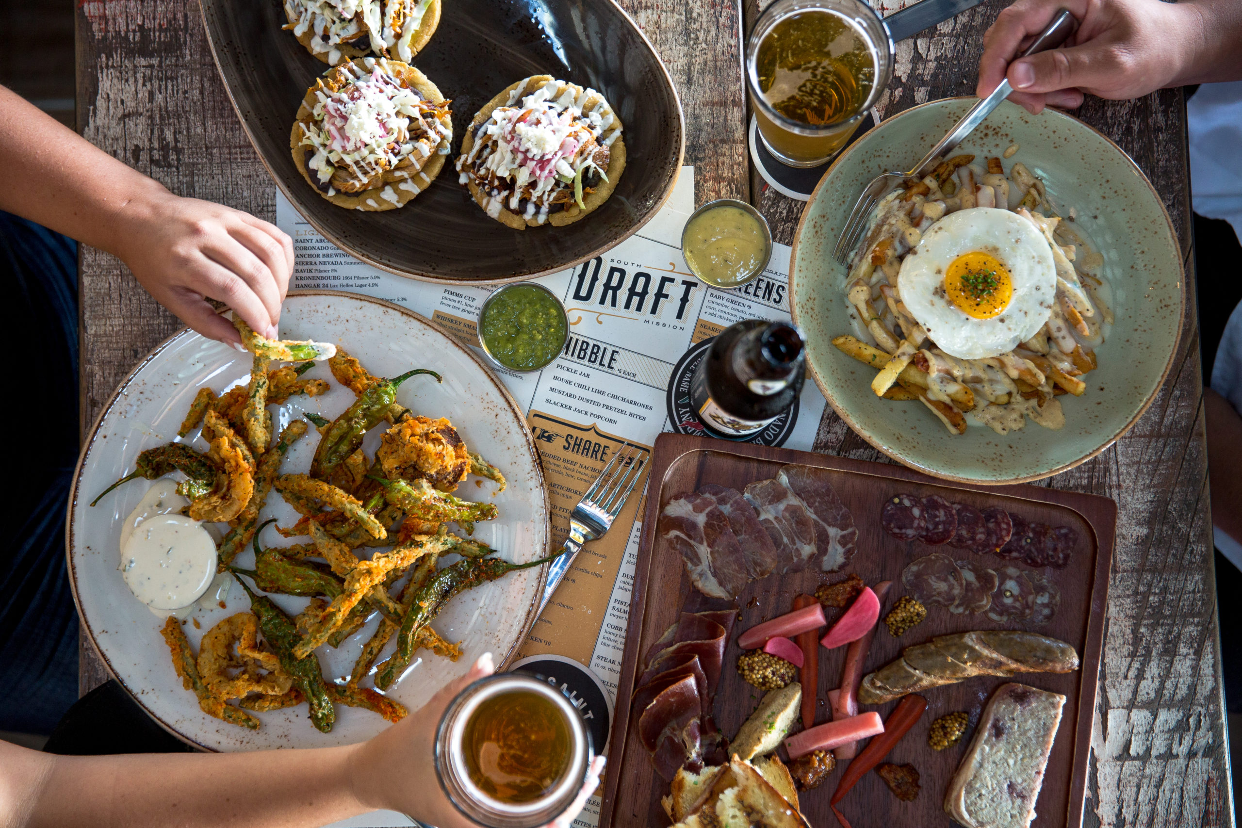 Overview shot of table at Draft with fries and shishito pepper and hands grabbing beers and silverware