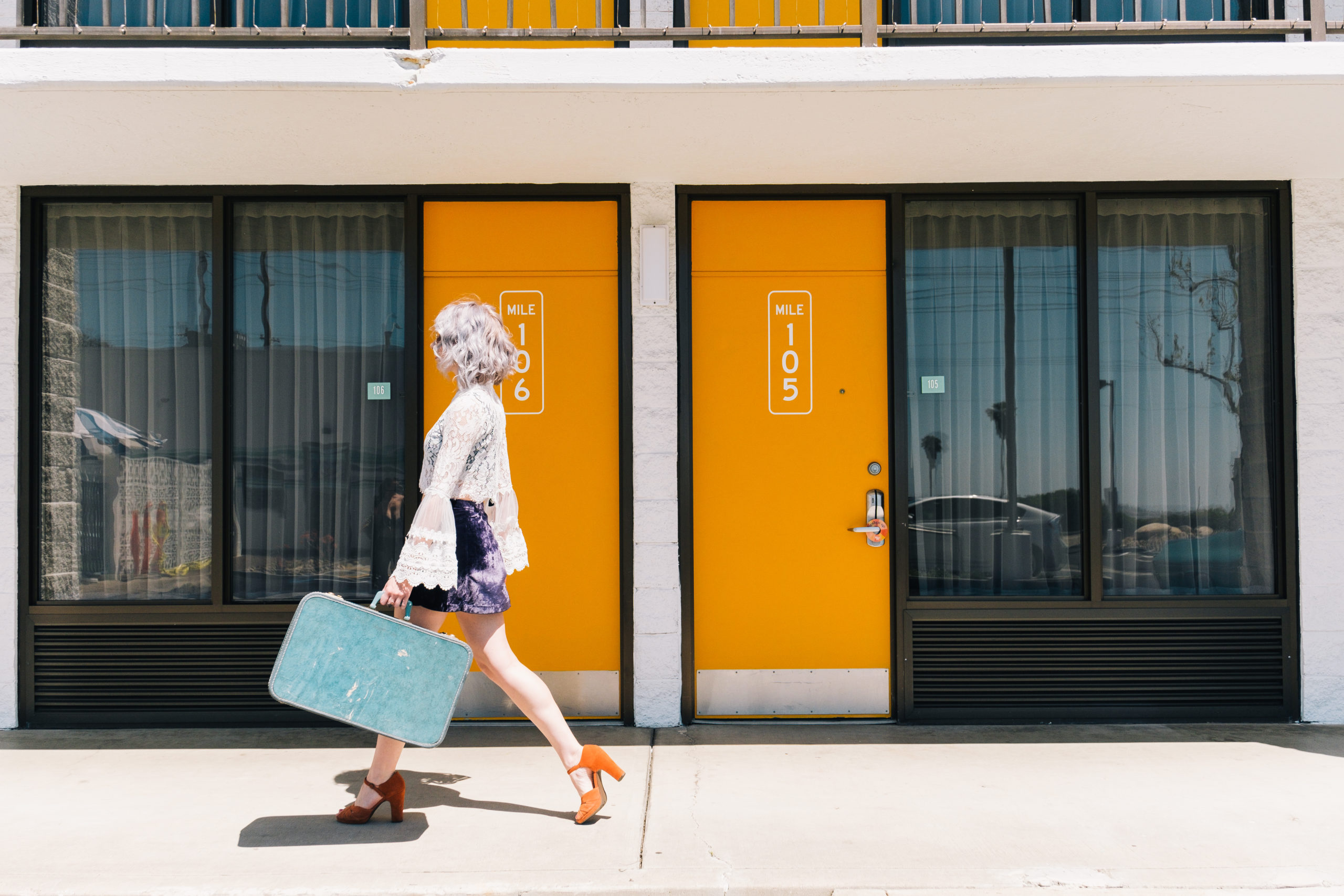 Woman walking with suitcase in front of yellow mile marker doors at Rambler Motel