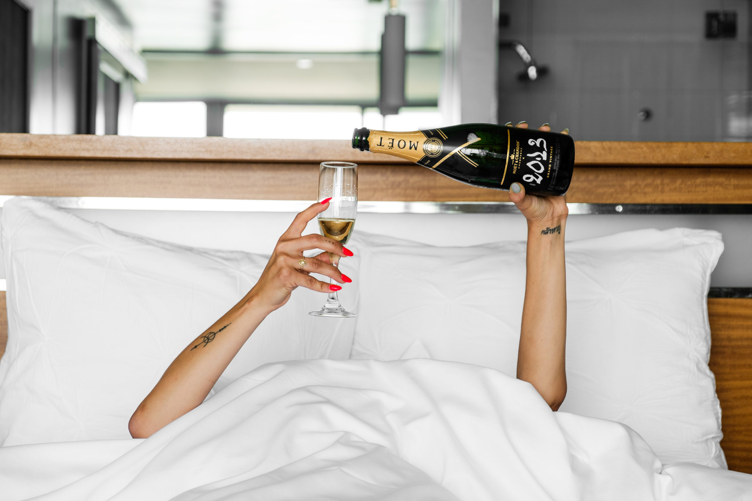 Hands popping out of the bed sheets pouring a bottle of champagne in one hand and holding a glass in the other