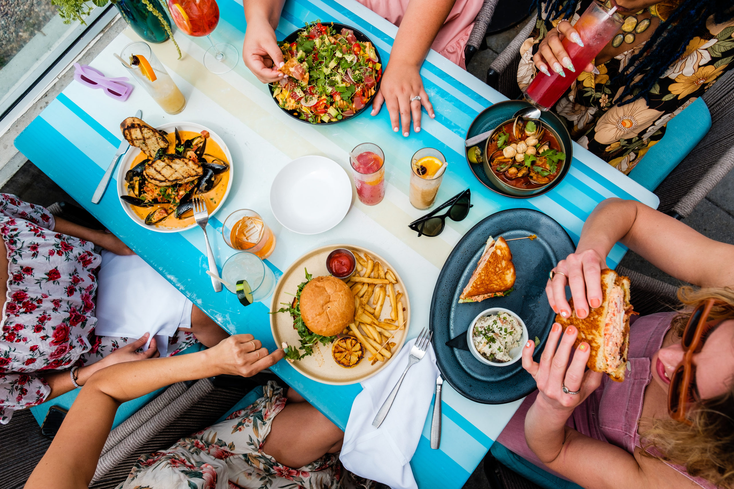 Birds eye view perspective lated table with a burger and fries and mussels with hands holding cocktails and heating sandwiches