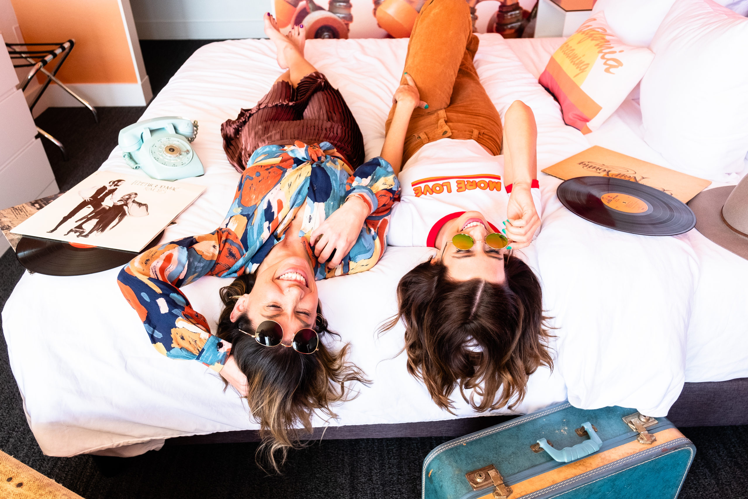 Two girls in sunglasses laying on a bed surrounded by records and suitcases smiling