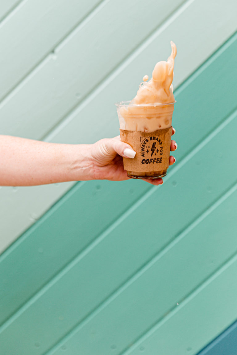 Hand holding a Rambler Coffee to go cup that is splashing its contents in front of a teal striped wall background