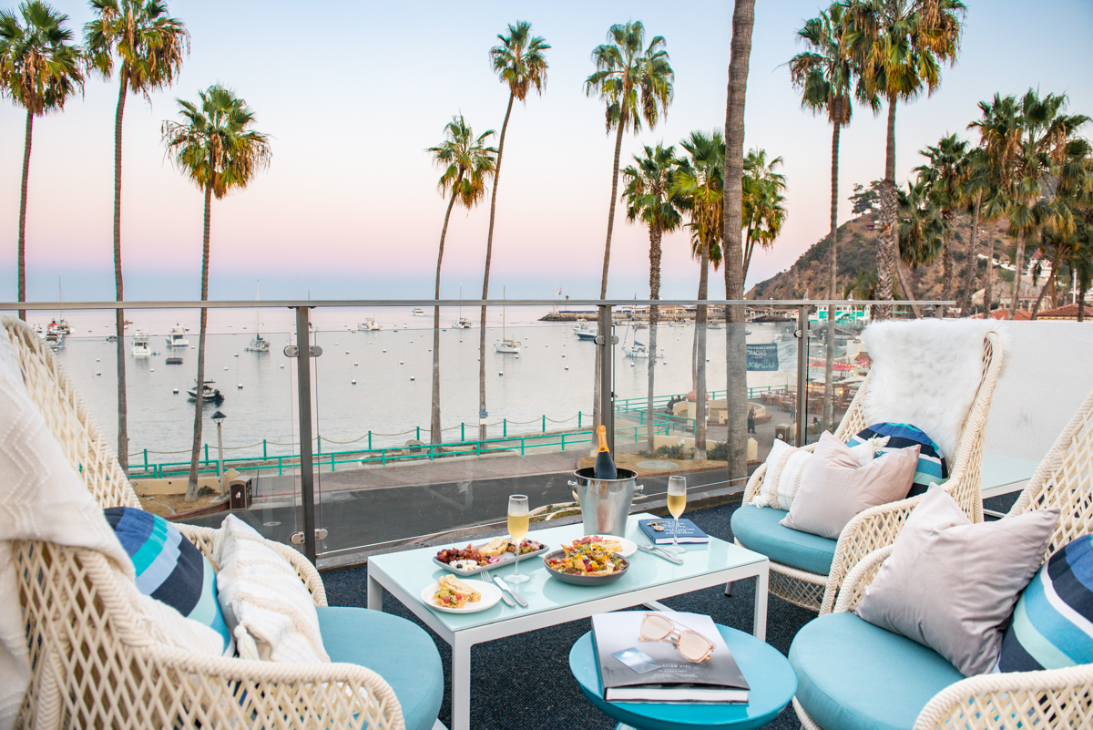 Plated meal with a bottle of champagne on the deck of Bellanca Hotel looking out onto the Avalon bay with boats and palm trees