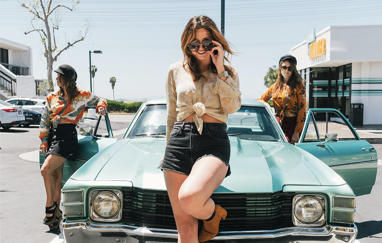 Girl in cut off shorts leaning on old school teal sports car in the Rambler Motel parking lots with two other girls standing near the doors
